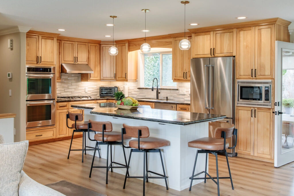 Open-concept luxury kitchen with custom wood cabinets, white island, dark granite countertop, stainless steel appliances, and tan leather barstools.