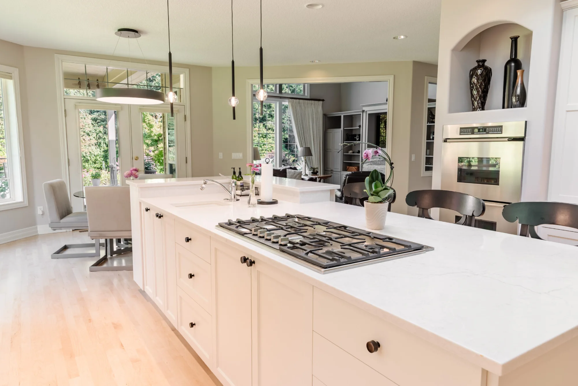 A beautifully remodeled luxury kitchen features a white quartz island with an integrated gas cooktop, white cabinetry, stainless steel appliances, and light hardwood floors.