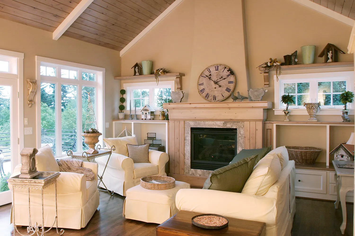 Warm living room with a stone-surround fireplace, wood-beam vaulted ceiling, slipcovered armchairs, and a large decorative wall clock.