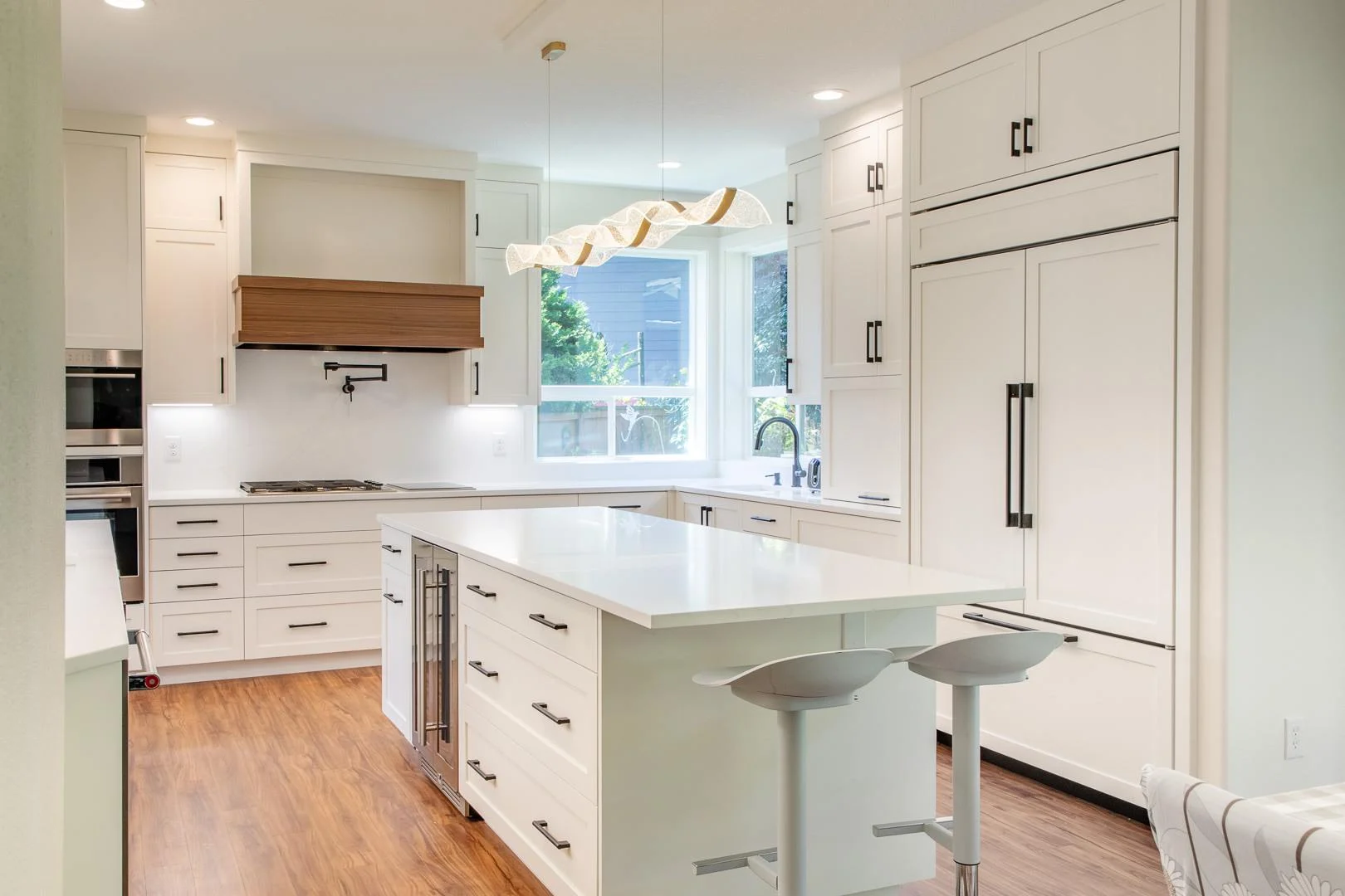 Contemporary white kitchen featuring a waterfall-edge island, wood vent hood, and paneled refrigerator, built by Black Cat Design Build LLC.