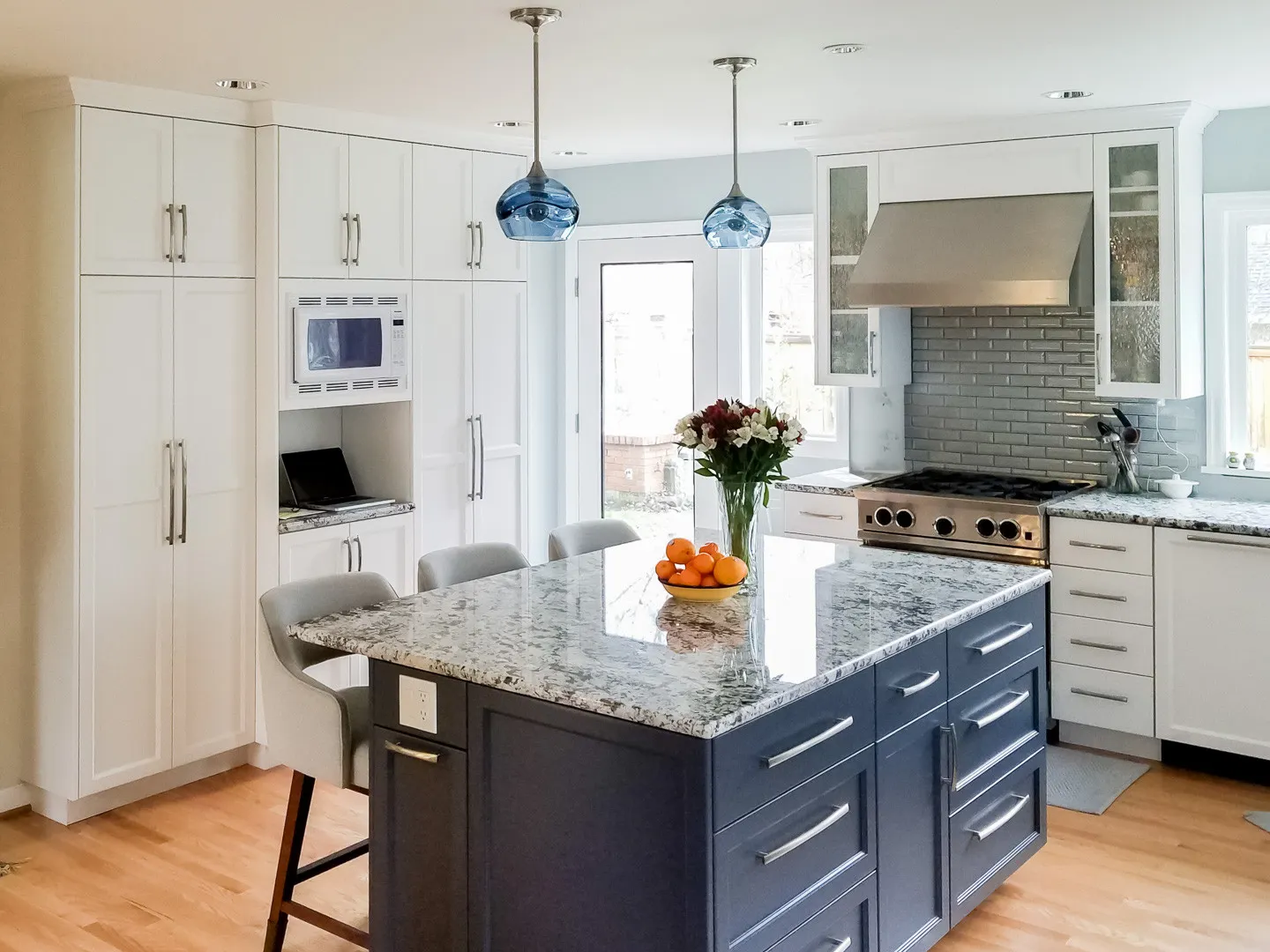 Modern kitchen with white shaker cabinets, a navy granite-topped island, blue glass pendant lights, and a stainless steel range.