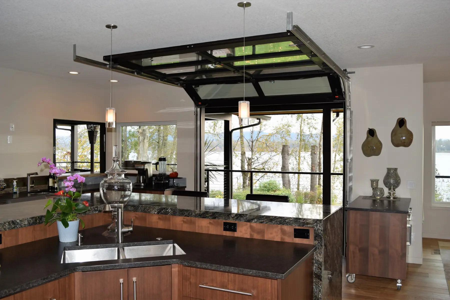 Modern kitchen with a lift-up glass garage door opening to a lake view, wood cabinetry, and dark granite countertops.
