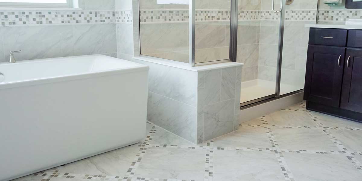 Modern bathroom featuring light marble tile flooring with patterned accent borders next to a white bathtub and glass shower.