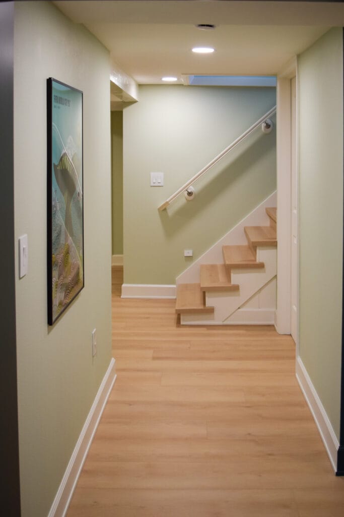 Light-colored luxury vinyl plank flooring in a remodeled basement hallway, reflecting light and brightening the staircase transition.